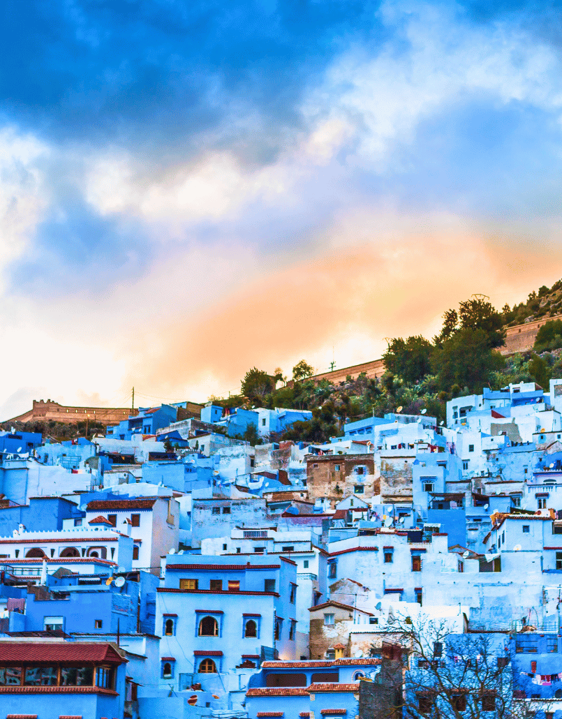 A hillside city with houses painted in shades of blue, under a colorful sunset sky.