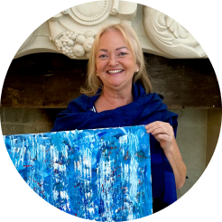 Older woman with gray hair smiling, holding a colorful abstract painting in front of a fireplace.
