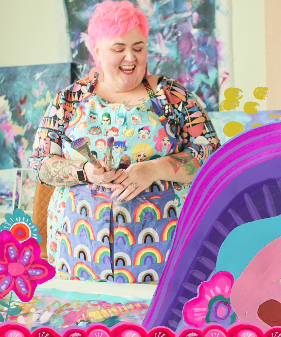 A woman with pink hair, wearing a colorful apron with cartoon faces and rainbows, is smiling and applying paint to a large canvas with a paintbrush. The background features vibrant, abstract artwork, and there are colorful paper or fabric flowers and decorations on the table in front of her.