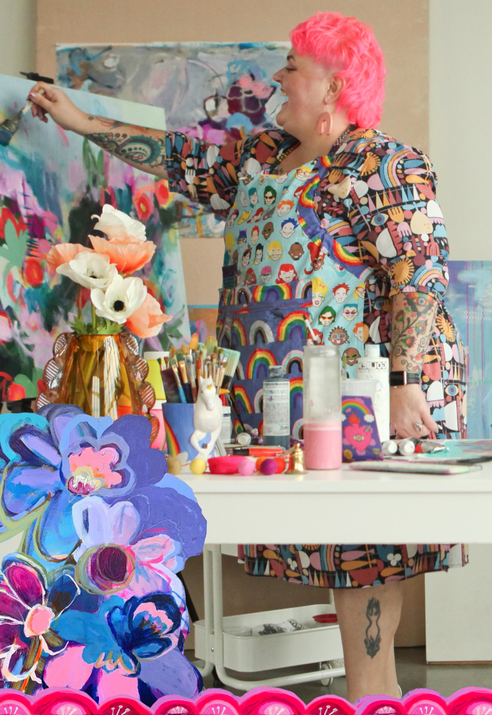 A woman with pink curly hair, colorful clothing, and tattoos is painting an abstract floral artwork on a canvas. She is wearing a patterned dress and a colorful apron with cartoon faces and rainbows. There are art supplies, including brushes, paint bottles, and a unicorn figurine, on the table in front of her.
