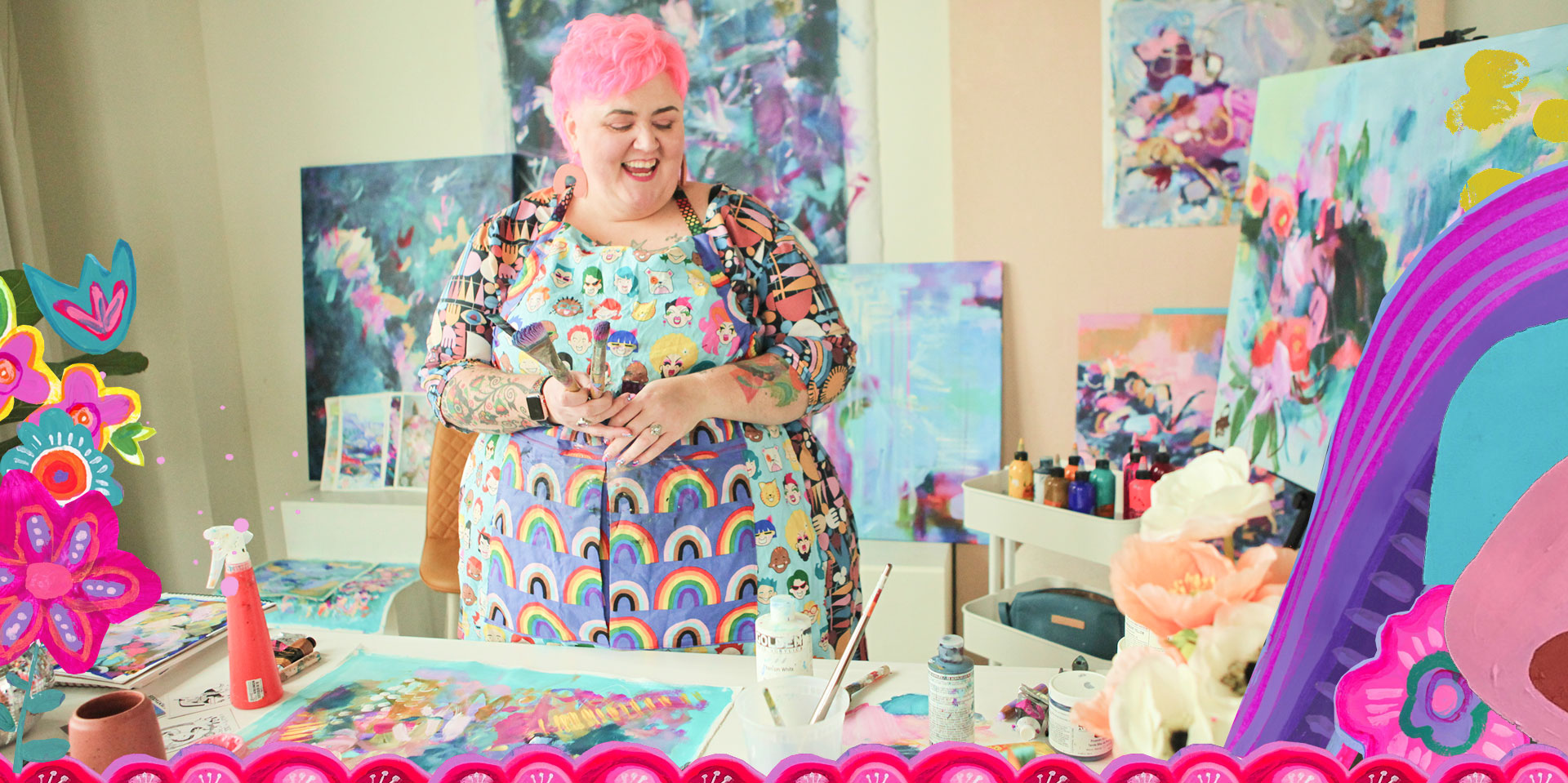 A woman with pink hair, wearing a colorful apron with rainbow and cartoon faces, is smiling while holding paintbrushes in her hand, standing in an art studio surrounded by vibrant paintings and art supplies.