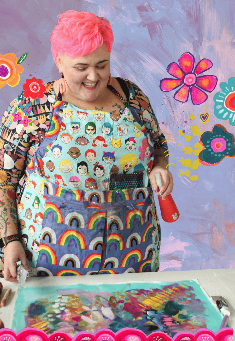 A woman with pink hair, wearing a colorful apron with rainbow and emoji patterns, is smiling in a vibrant art studio with painted flowers and abstract patterns on the wall. She is holding a red spray paint can, working on a colorful canvas.