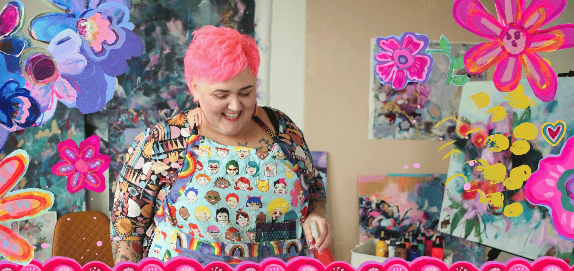 A woman with short pink hair smiling, wearing a colorful apron with various cartoon faces, in a vibrant art studio with colorful paintings and paper flowers surrounding her.