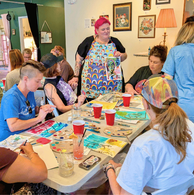 Group of young people participating in an art class, painting with various colors, led by an instructor with pink hair and a colorful apron, in a cozy, decorated art studio or cafe.
