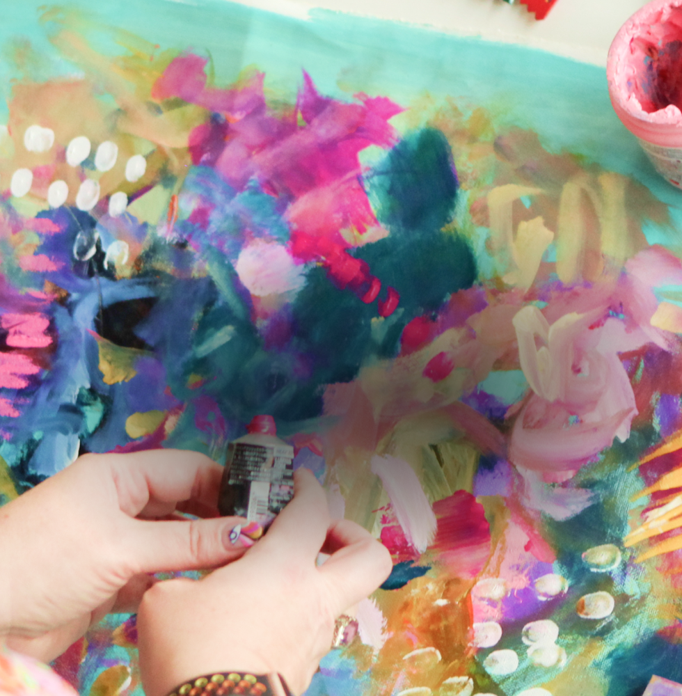 Close-up of an abstract, colorful painting being created with a person's hand holding a small tube of paint, on a surface filled with vibrant, expressive brushstrokes and colors.