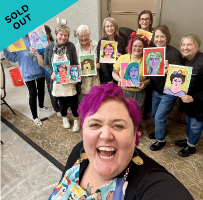 Group of women at an art event, holding colorful portrait paintings of women with various hairstyles and accessories, with one woman in the foreground taking a selfie. The image has a sold-out banner in the top left corner.