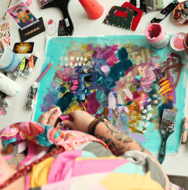 Top-down view of an artist working on a colorful abstract painting surrounded by art supplies, including paint bottles, brushes, and a palette knife.