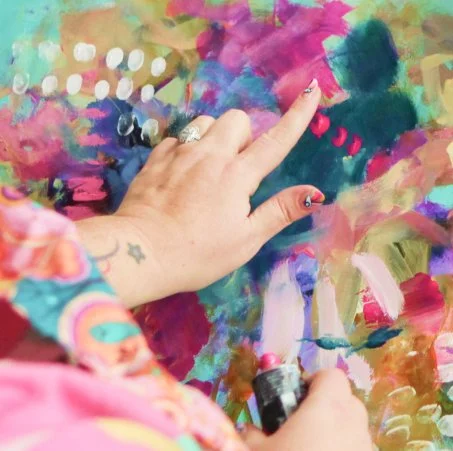A person's hand with decorated nails and rings is touching a colorful abstract painting with various bright colors and patterns.