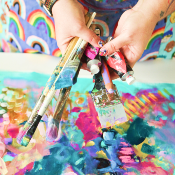 Person holding multiple paintbrushes with colorful abstract painting in background.