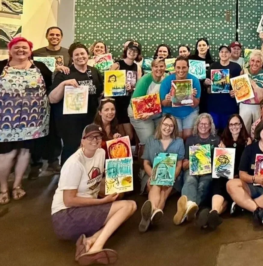 Group of people at an art event holding colorful paintings, smiling, with a green polka dot background.