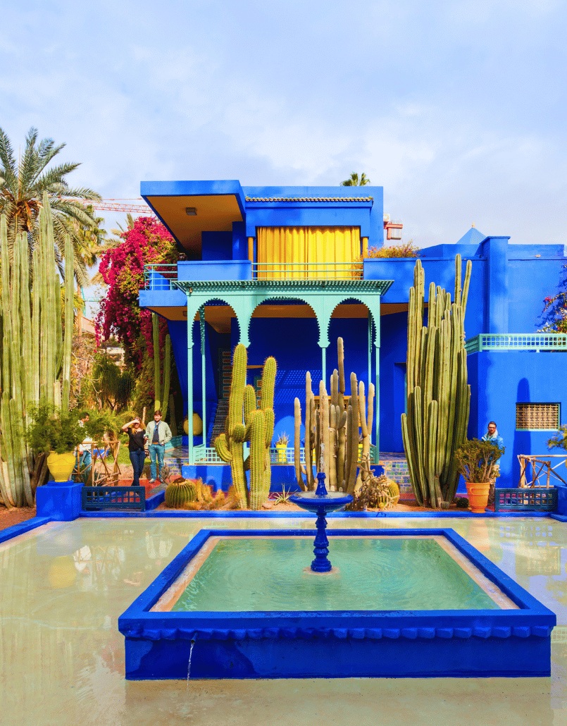 Colorful building with blue walls and yellow curtains, surrounded by tall cacti, with a rectangular fountain in the foreground, and people walking near the building.