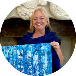 A smiling woman with gray hair holding an abstract blue painting in front of a fireplace.