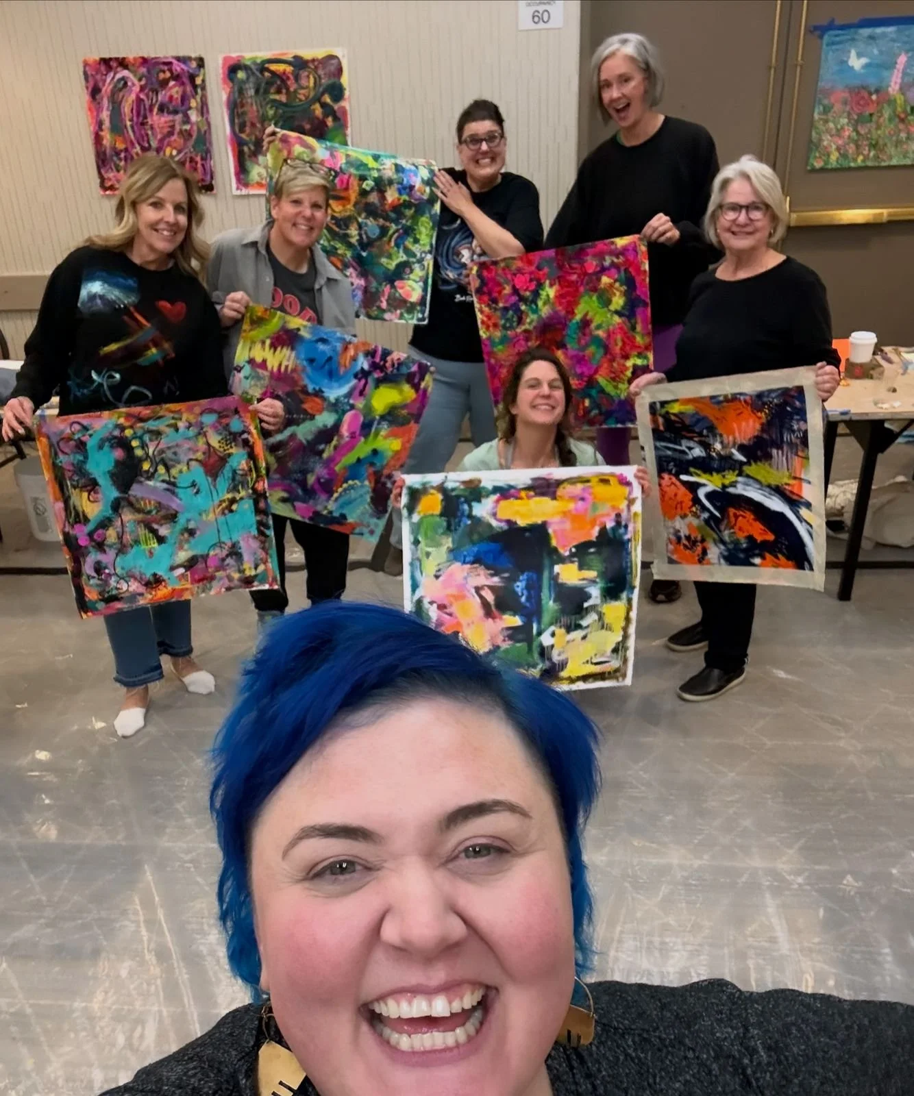 Group of women holding colorful abstract paintings in an art class or gallery setting, with paintings hanging on the wall behind them and one woman taking a selfie in the foreground.