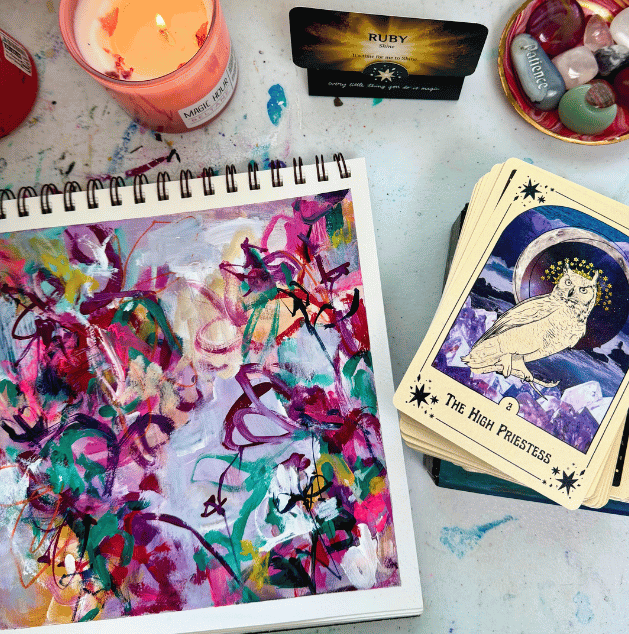 An art desk with a colorful abstract painting, a burning candle, a tarot card deck with 'The High Priestess' card, a deck of crystals, and a black card with 'RUBY' written on it.