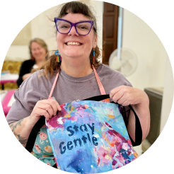 Woman smiling and holding a colorful tote bag with the words 'Stay Gentle' printed on it, in a room with other people in the background.