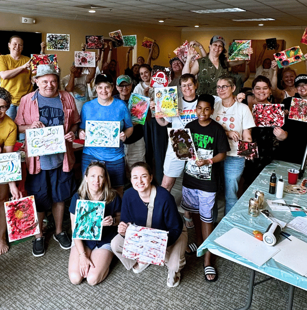 Group of people at an art event showing paintings they've created, some seated or kneeling, others standing, smiling, and holding colorful abstract artwork.