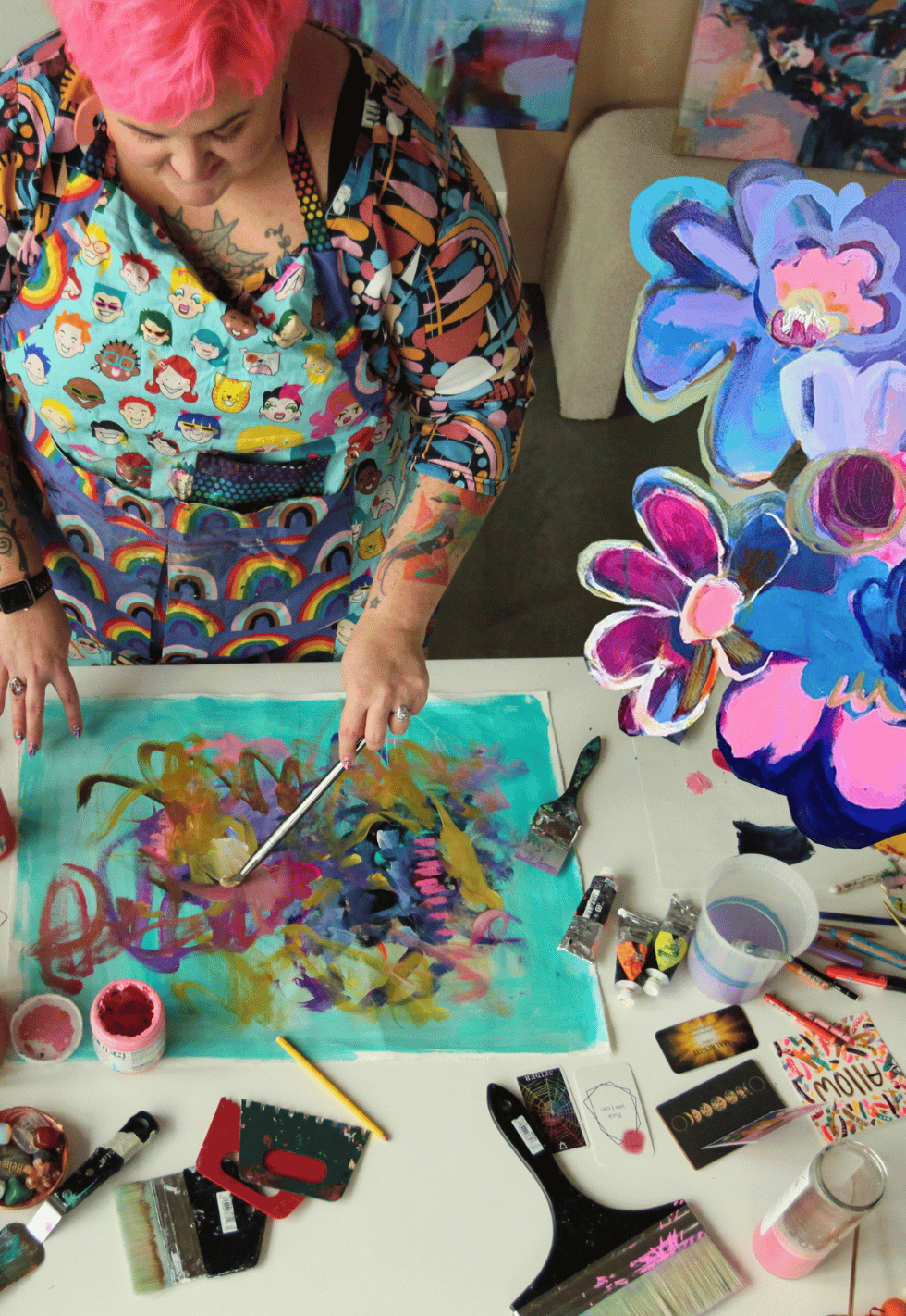 A woman with pink hair, wearing a colorful apron with smiley faces, is painting a colorful abstract picture on a table. Art supplies like paints, brushes, and cards are arranged on the table.