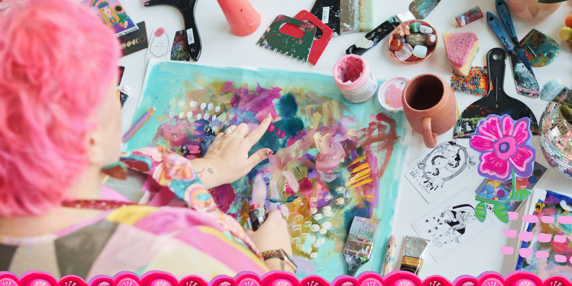 A person with pink hair creating abstract art on a colorful paper at a cluttered table filled with paints, brushes, mugs, and decorative items.