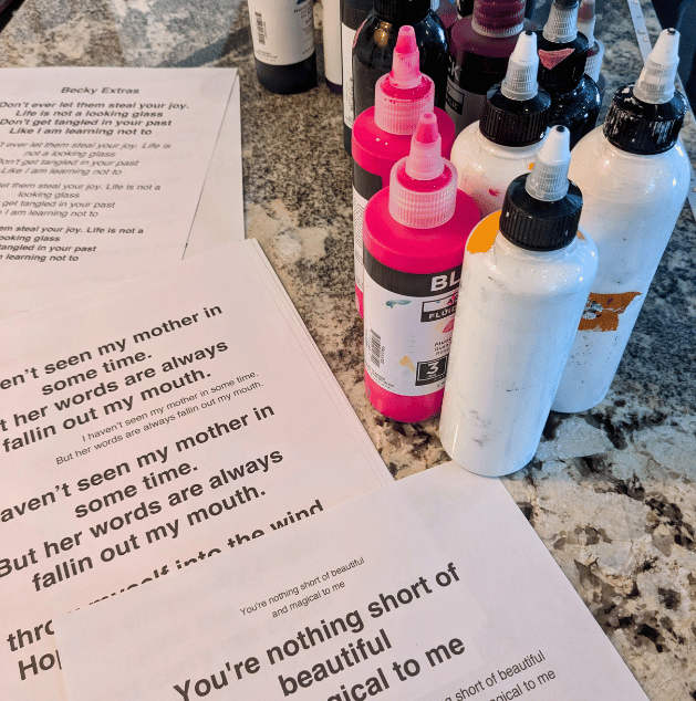Assorted bottles of acrylic paint, including pink, white, and black, on a kitchen counter next to printed song lyrics and a sheet of paper.