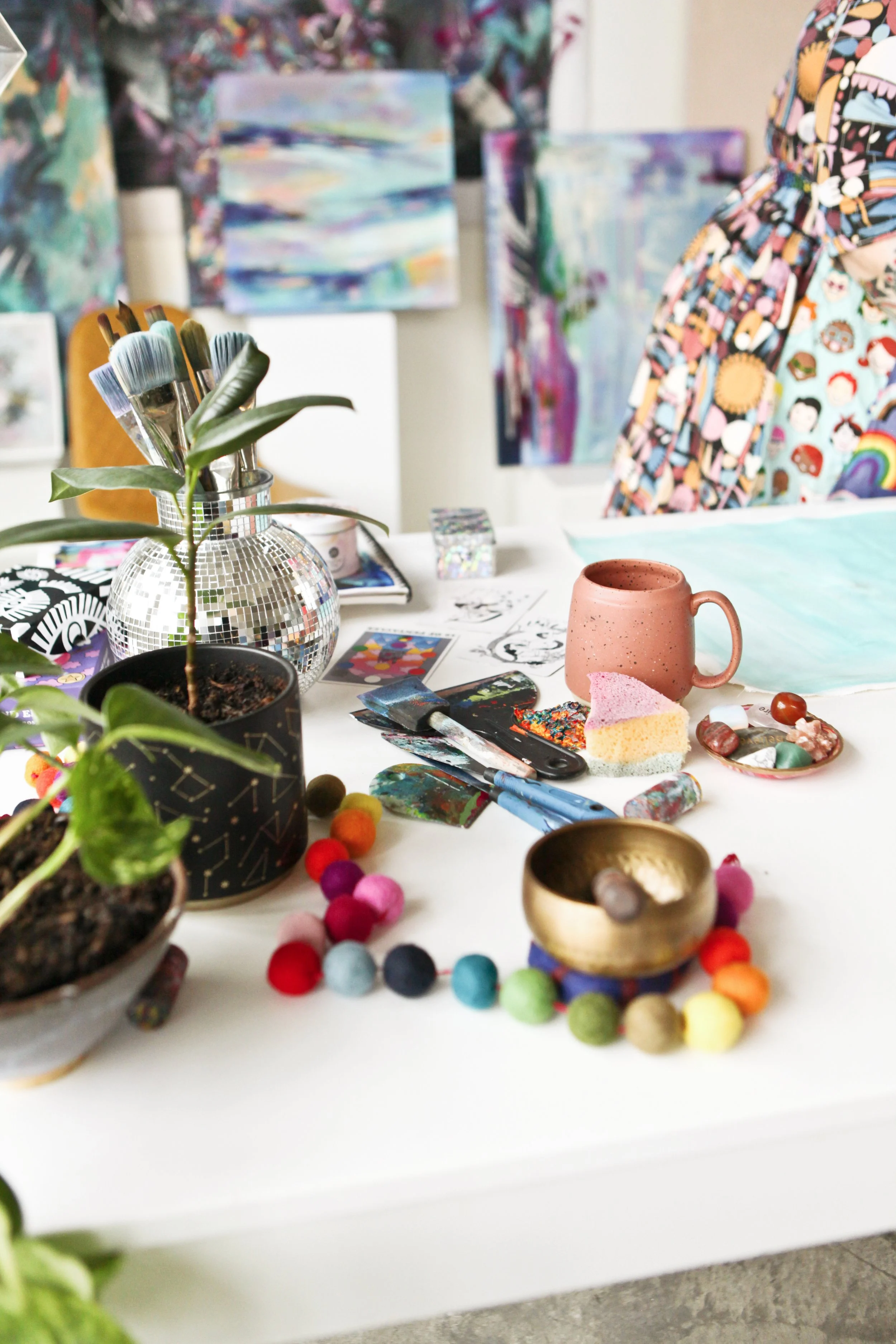 A collection of small potted plants, art supplies including brushes, a spatula, and paint, colorful felt balls, a ceramic mug, and abstract artworks in the background.