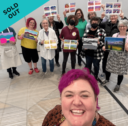 Group of women at an art show holding landscape paintings, with one woman in the foreground taking a selfie, all smiling and some wearing masks.