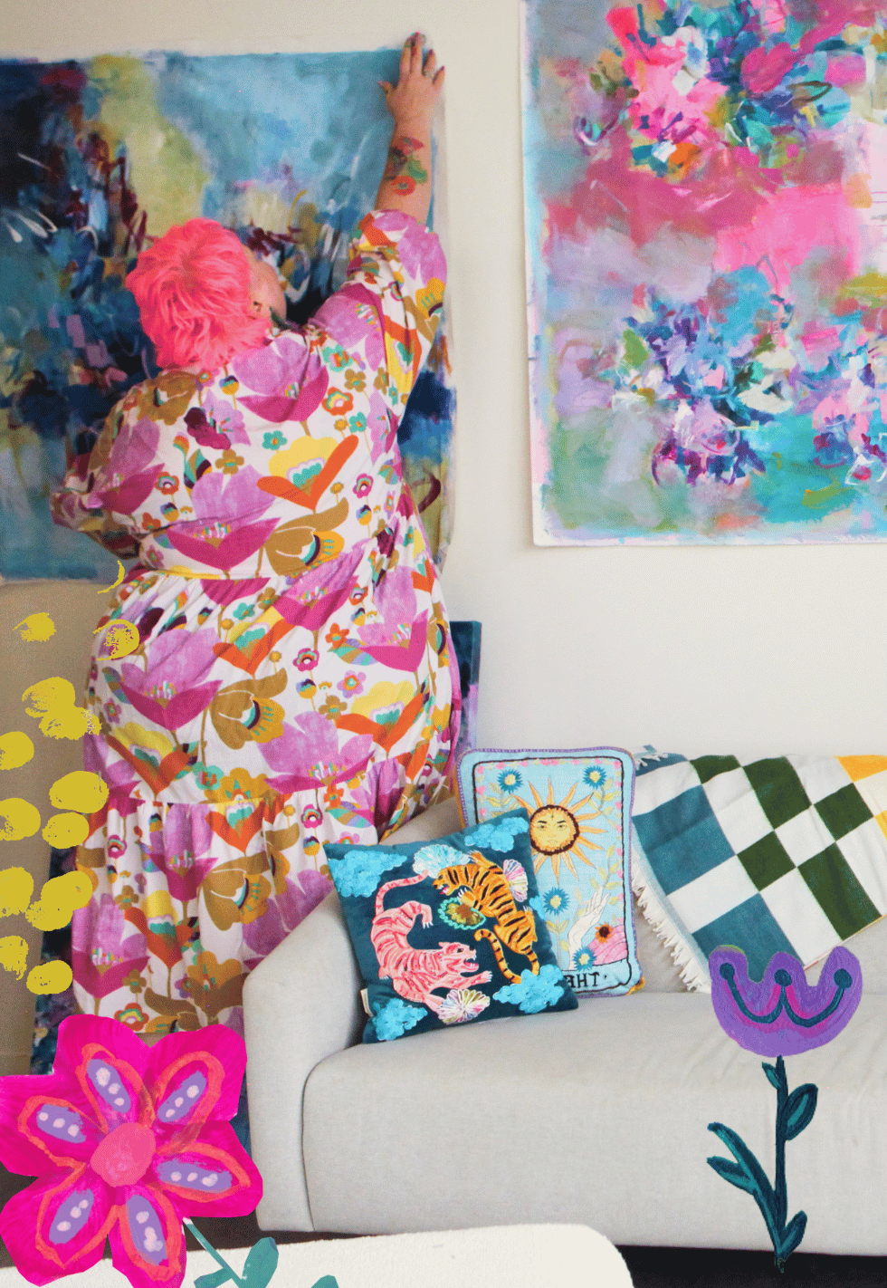 A person with pink hair hanging a blue painting in an art gallery, surrounded by colorful floral paintings and cushions with tiger and sun designs on a beige sofa, with added illustrated flowers and yellow dots.