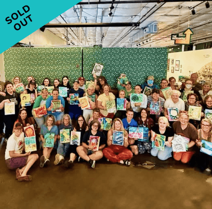 Group of people holding up colorful paintings or artwork in an indoor space with green walls and overhead lighting.