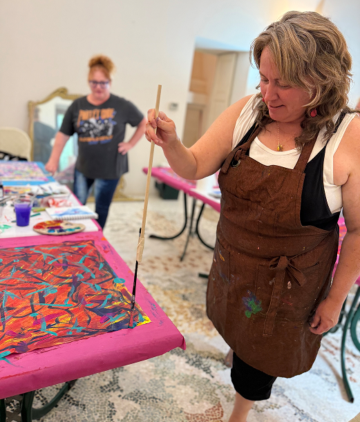 Woman painting with a long brush on a colorful canvas while another woman stands nearby in a room with tables covered in art supplies.