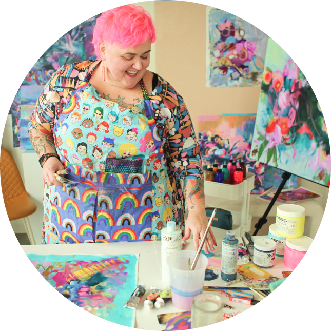 Becky Joan Springer - an artist with pink hair wearing a colorful apron with rainbow patterns, working in a brightly decorated art studio filled with paintings, paints, and art supplies.