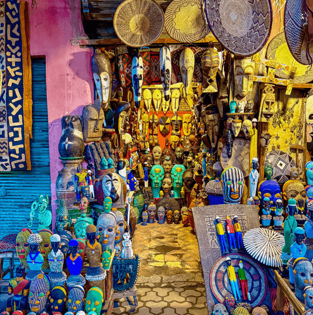 Street vendor stall displaying colorful African masks and sculptures, with some woven baskets hanging overhead.