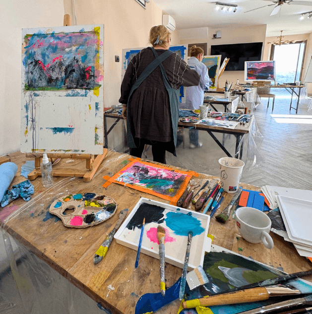 Art studio with paintings, brushes, palettes, and art supplies on tables. Two women are painting, with one facing away and the other to the side. Large windows let in natural light.
