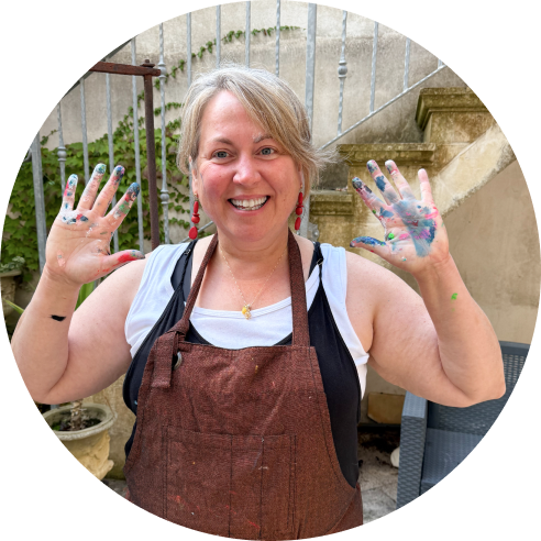 A smiling woman with paint on her hands, wearing a brown apron and earrings, celebrating her painted hands outdoors.
