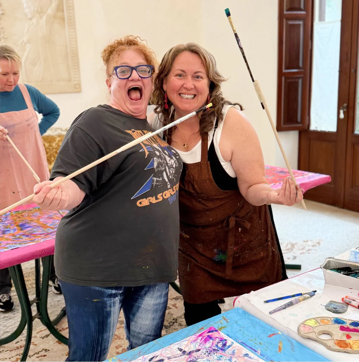 Two women smiling together while holding paintbrushes, in a room with painting supplies and colorful artwork, during a painting class or art activity.