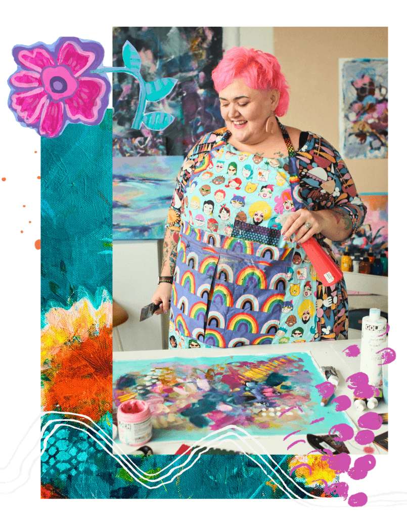 A woman with pink hair wearing a colorful apron with rainbow and emoji patterns, standing at an art studio table with paint supplies and colorful artwork in the background.