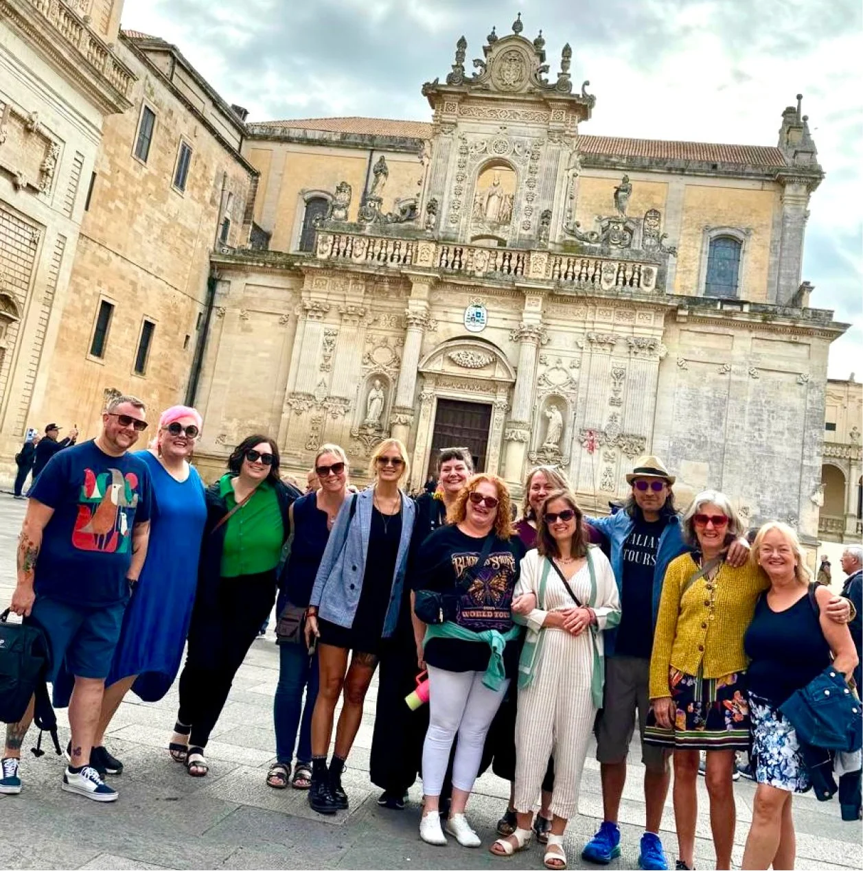 A group of eleven tourists smiling and posing in front of an ornate historic building with detailed carvings, statues, and a large wooden door, on a cloudy day.