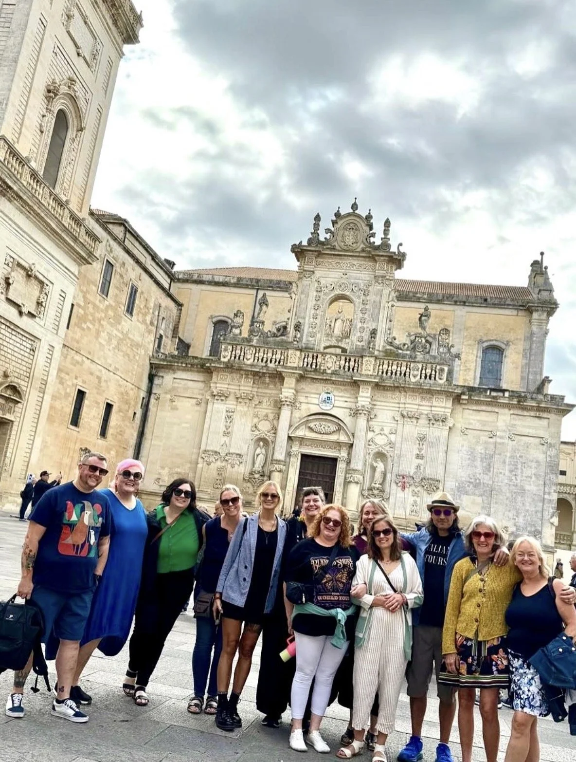 A group of diverse tourists smiles and poses for a photo in front of a historic European church with ornate stone architecture, statues, and a balcony. The sky is cloudy, and some people in the background are walking around.