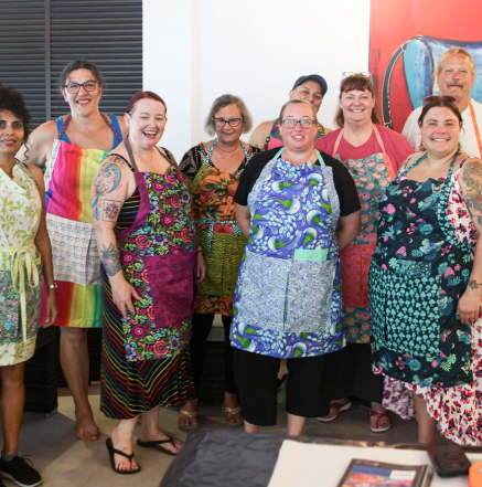 Group of women standing together in colorful aprons and casual clothing, smiling at the camera in an indoor setting.