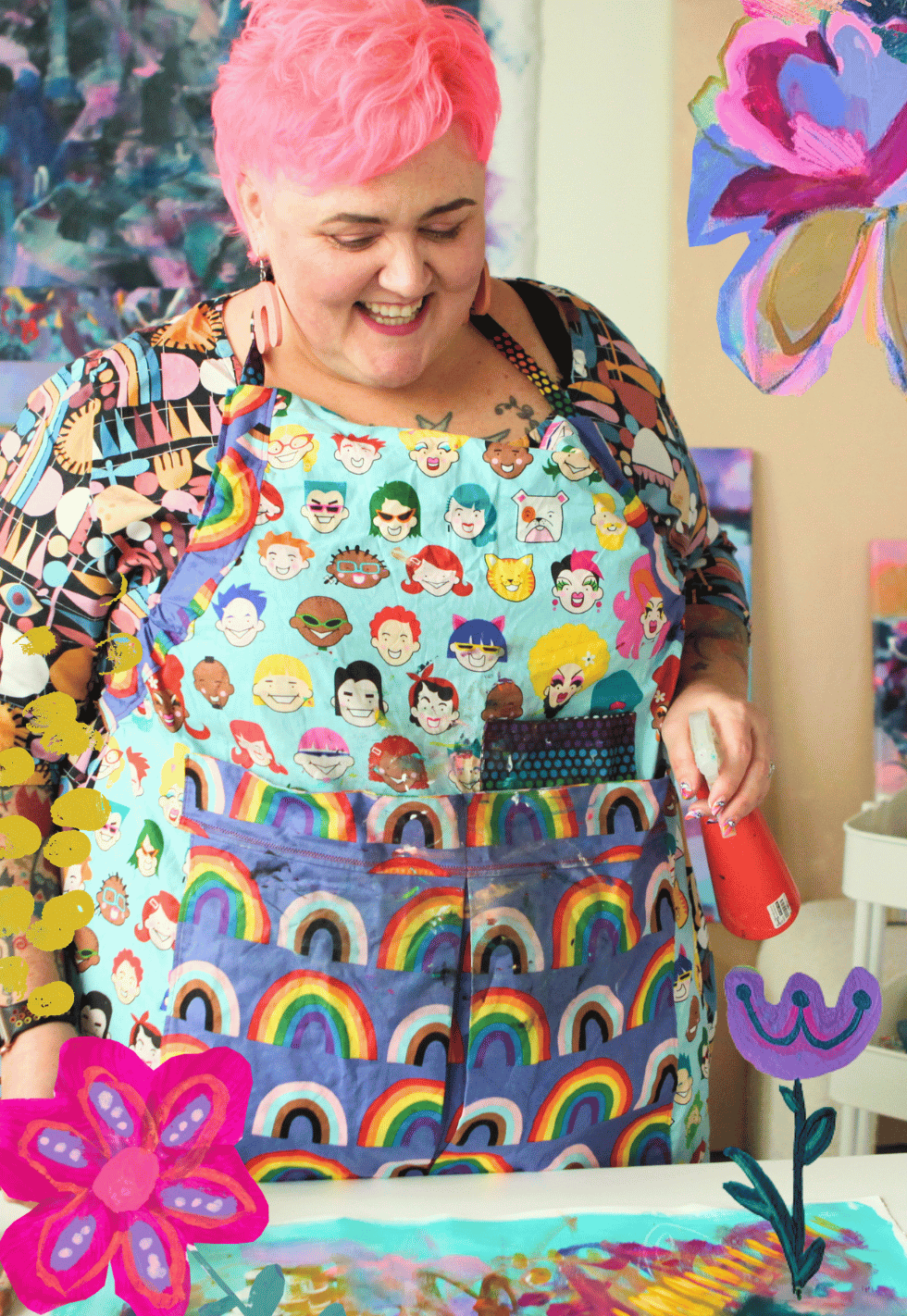 A woman with pink hair is smiling, wearing a colorful apron with cartoon faces and rainbow patterns, standing in front of a table with colorful painted flowers and artwork.