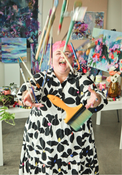 A woman with pink hair wearing a black and white patterned dress, smiling and throwing paintbrushes in the air inside an art studio with colorful paintings and art supplies in the background.