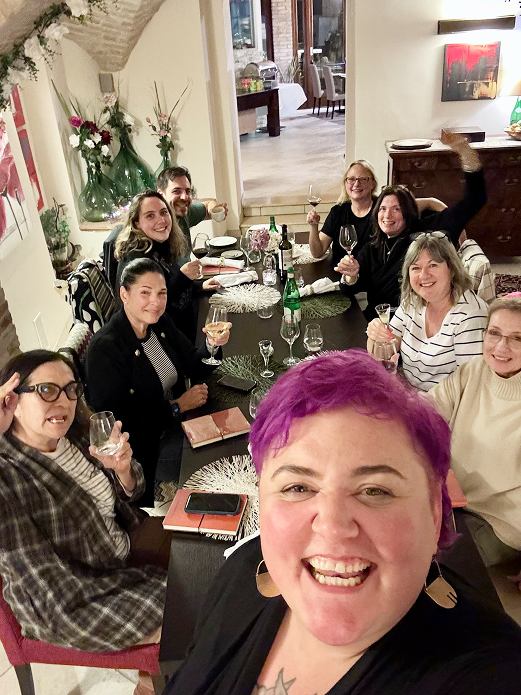 A group of nine people gathered around a dining table, smiling and enjoying drinks at a dinner party in a cozy, decorated home interior.