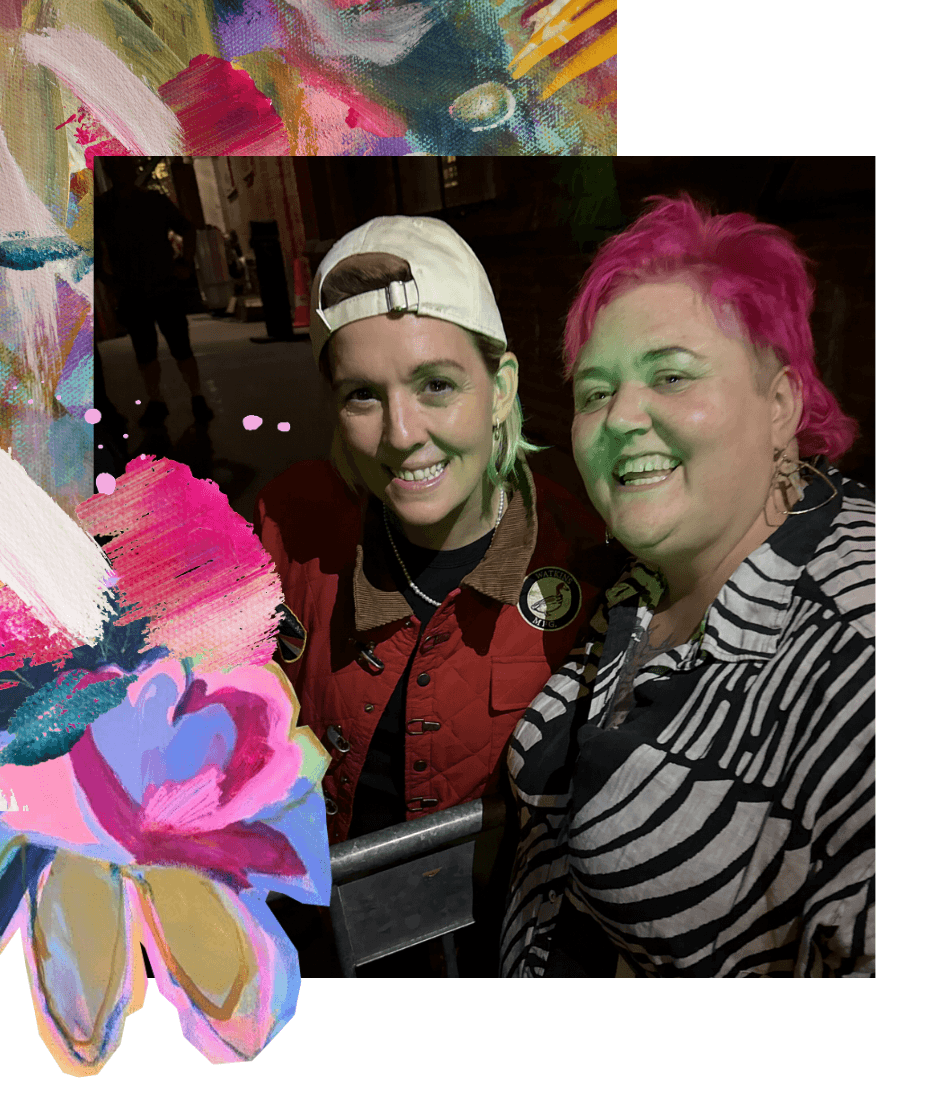 Two women smiling for a photo at night. One with pink hair and the other wearing a beige cap and red jacket, with colorful artistic elements and flowers surrounding the photo.