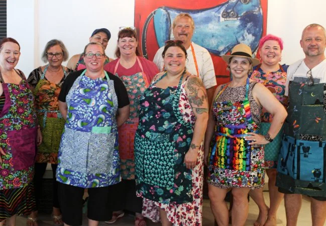 Group of people wearing colorful aprons, smiling, standing indoors with a large blue canon painting in the background.