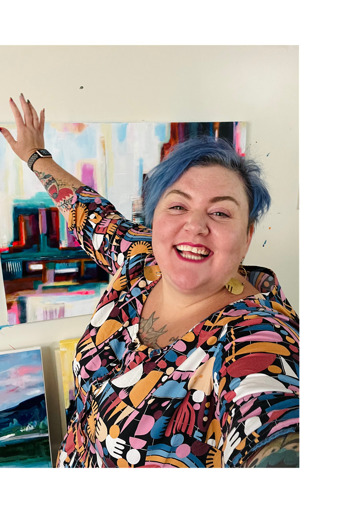 Smiling woman with short blue hair, wearing a colorful patterned dress and gold earrings, standing in front of an abstract art painting indoors.