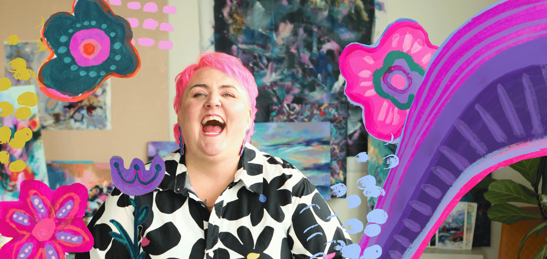 A woman with short pink hair, laughing with her mouth open, sitting in an art studio. She wears a black and white patterned shirt. The background includes colorful paintings and drawing of bright pink, purple, and blue flowers and abstract elements.