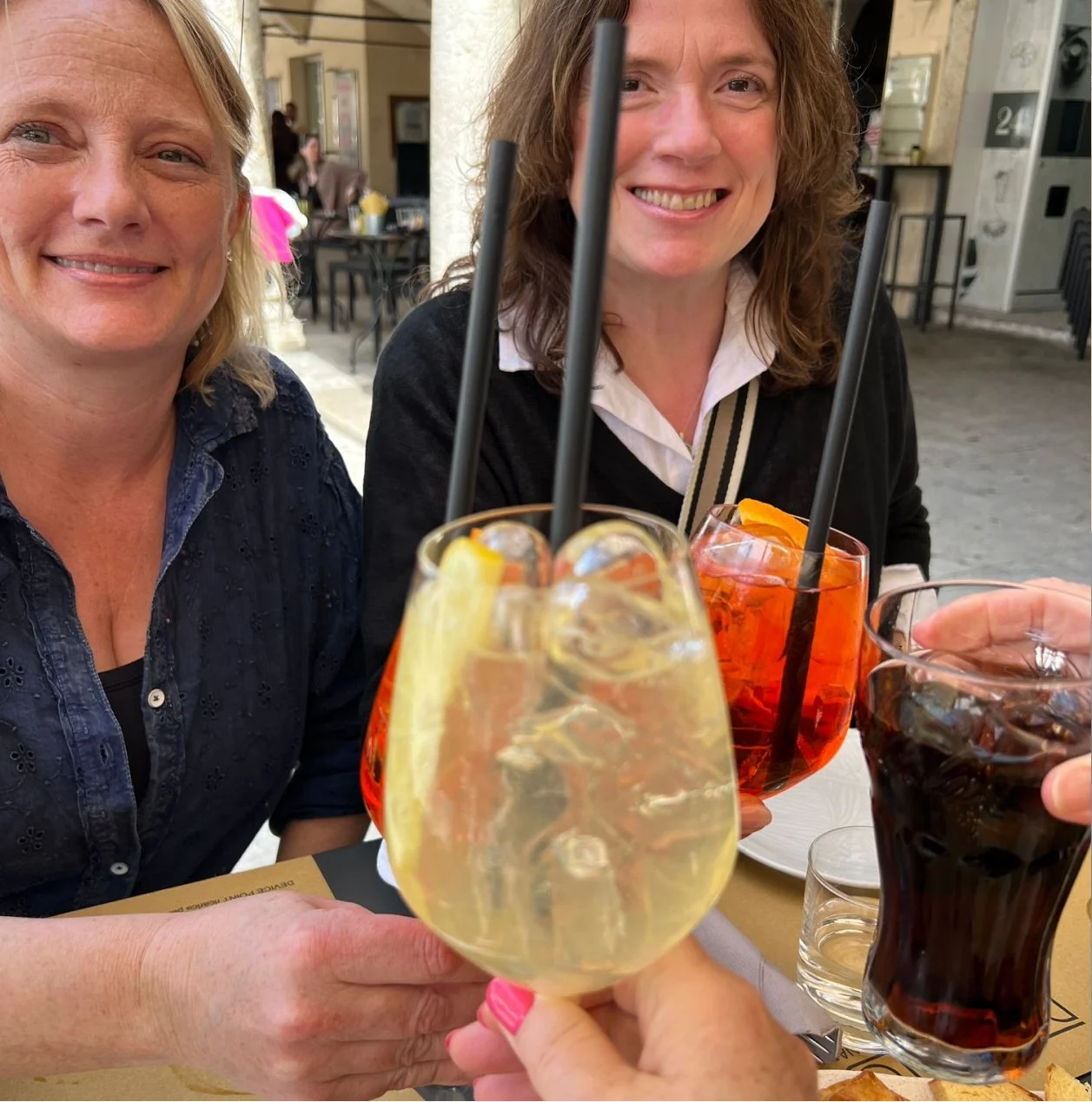 Two women smiling at a table with various colorful drinks, including cocktails with slices of lemon and orange, and a glass of soda, possibly during a celebration or casual gathering.