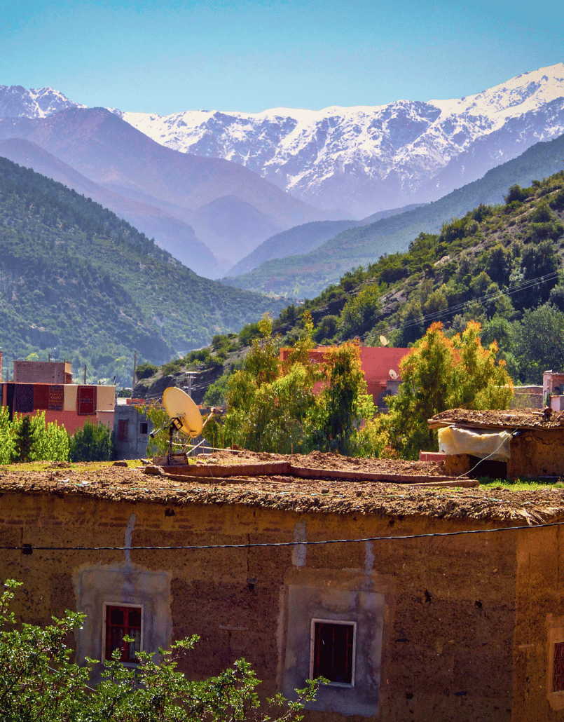 Mountain landscape with snow-capped peaks, green hills, and a small village with buildings and trees in the foreground.