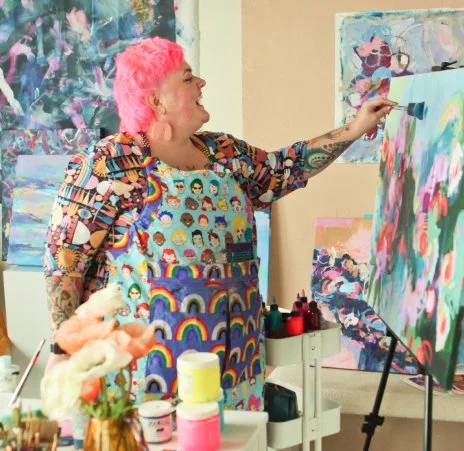 Becky Joan Springer with pink hair painting on a large canvas in an art studio.
