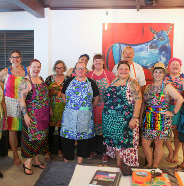 A group of ten people, mostly women, wearing colorful aprons and casual clothing, standing together indoors in front of a large red and blue cow painting.