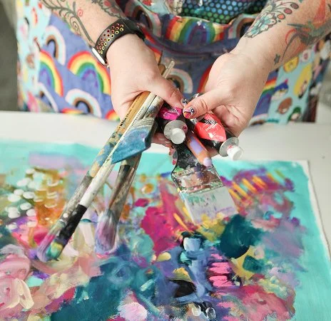 Person mixing vibrant acrylic paints with palettes and brushes on a colorful abstract artwork.