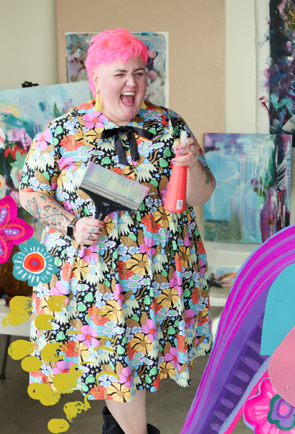 A woman with bright pink hair, wearing a colorful floral dress, is smiling and holding a paint scraper and a bottle of pink paint in her studio surrounded by vibrant abstract paintings.